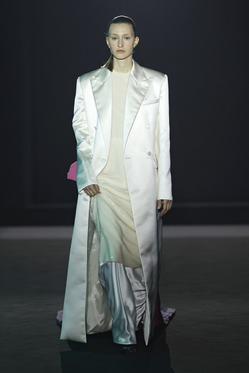 Modelo desfilando en la 080 Barcelona Fashion con un look total white donde destaca un abrigo extralargo en tejido satinado y vestido de lentejuelas del diseñador Juan Vidal 
