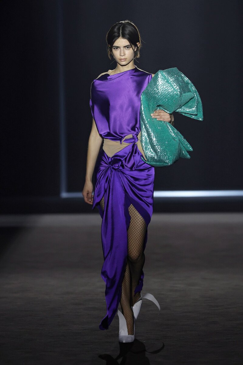Modelo desfilando en la 080 Barcelona Fashion con un look de Ernesto Nara compuesto de vestido en dos piezas y lazada en la cinturo de color violeta y maxibolso verde agua