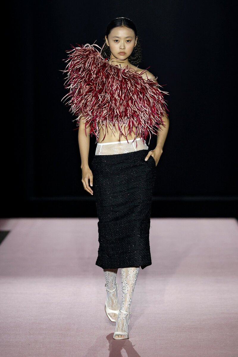 Modelo desfilando un look femenino de COMPTE SPAIN con un top asimétrico de plumas color borgogna y falta negra con cintura ancha blanca