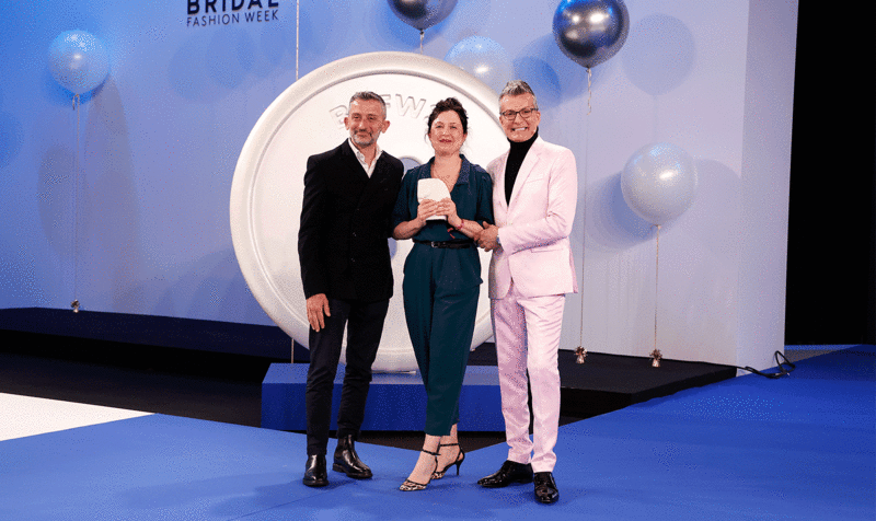Foto entrega de premios Barcelona Bridal & Fashion Awards con Andrea Marchesi a la izquierda, junto a la diseñadora premiada y el presentador de la gala delante de un fondo azul 