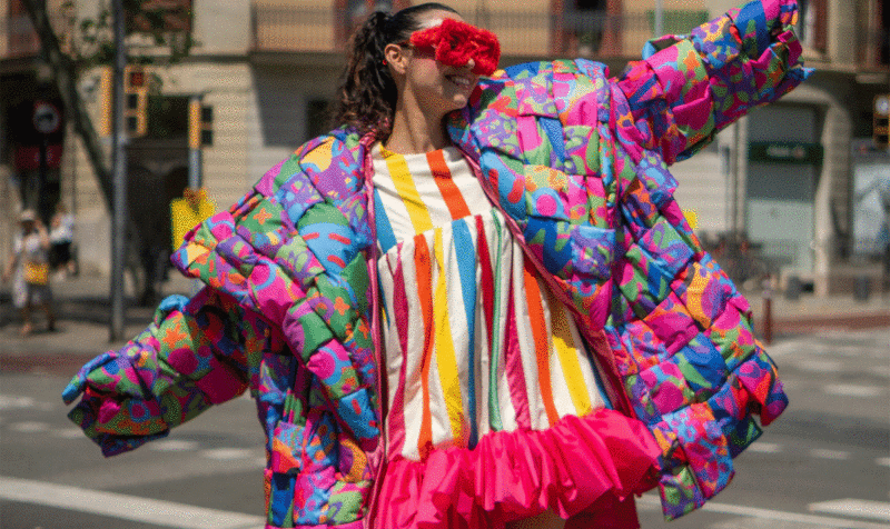 Una chica con grandes gafas rojas y un abrigo oversize tipo patchwork sobre un vestido de rayas de colores en expresión alegre en medio de la calle