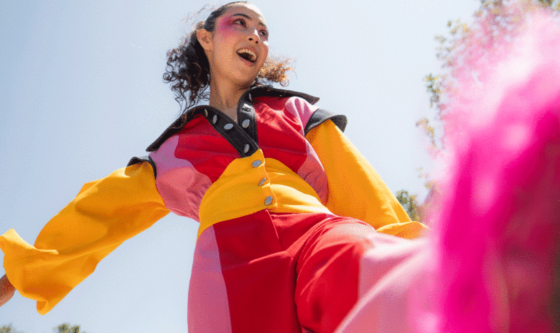 Plano contrapicado de una chica alegre dando una patada al aire. Viste un mono colorblock en tonos rojo, rosa, amarillo y negro con cuello camisero