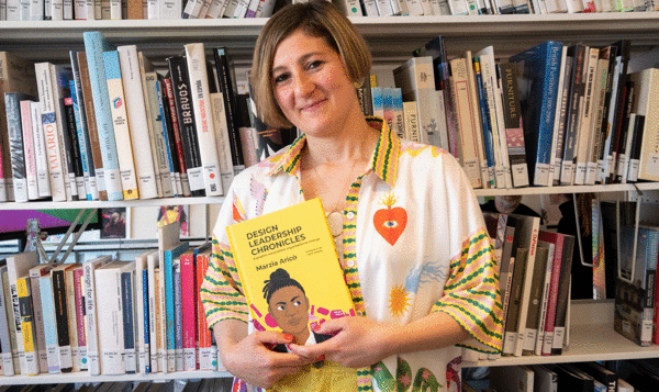 Una mujer rubia sostiene un libro amarillo titulado Design Leadership Chronicles delante de una gran estantería llena de libros
