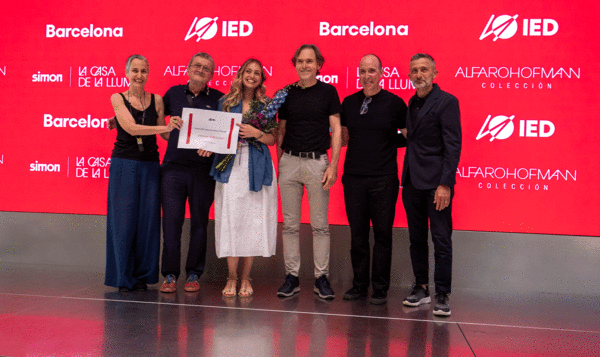 Un jurado de 5 personas entrega un diploma a una estudiante sobre un escenario gris con una pantalla con fondo rojo y logos IED Barcelona en el fondo