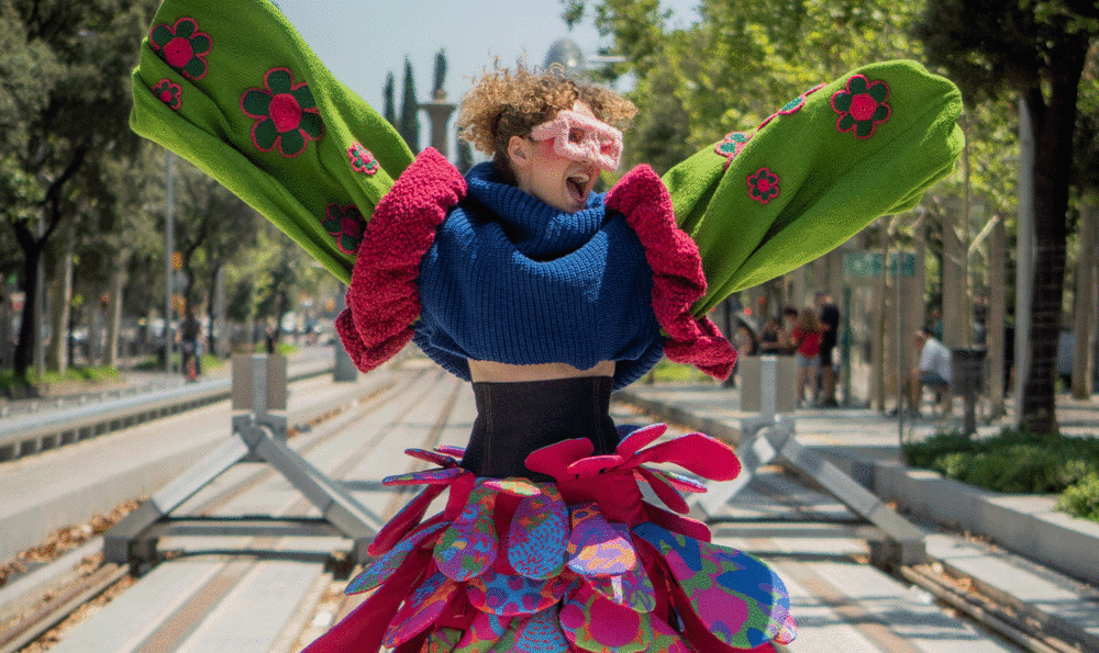 Una chica joven alegre con los brazos abiertos y expresión feliz. Viste gafas XXL y ropa muy colorida frente a las vías del tramvía